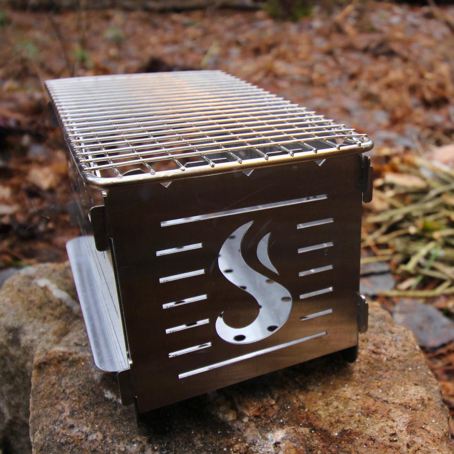 CAMPKITCHEN Twig Stove. Stove is on an angle showcasing the back end complete with the CAMPKITCHEN flame.