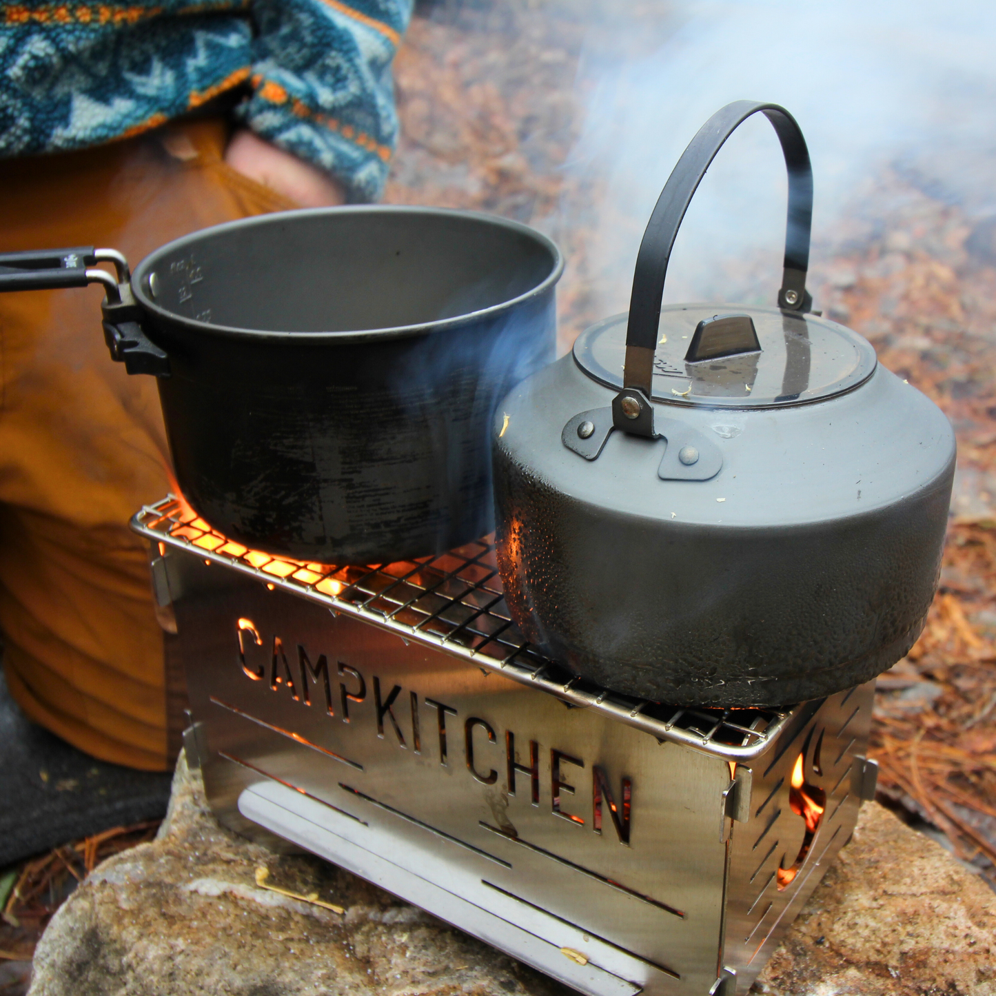 CAMPKITCHEN Twig Stove in action. Stove is lit with a pot and a kettle ready to rock. Flames light up the CAMPKITCHEN logo.