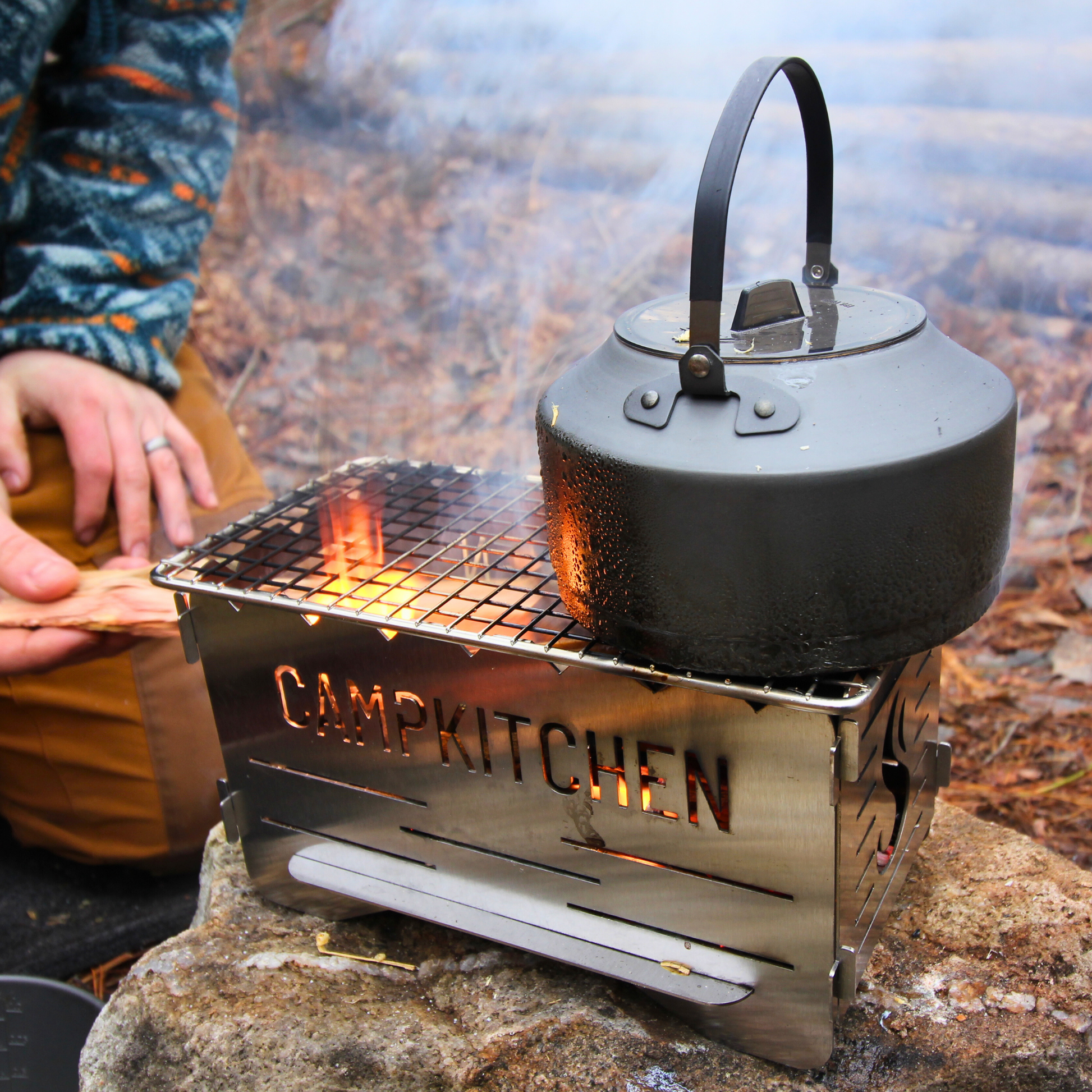 CAMPKITCHEN Twig Stove in action. Stove is lit with a teapot on the right side. Flames appear through the left side of the grate showcasing space to grill or to add another pot/pan.